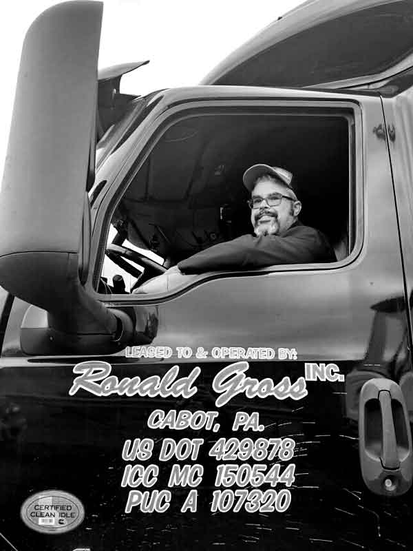 A Ronald Gross Inc. flatbed truck driver smiles form the seat of the cab in black and white.