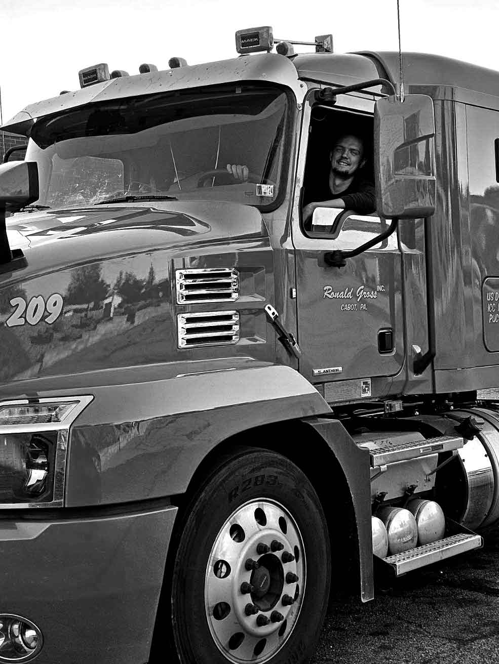 A black and white photo of a Ronald Gross Inc. flatbed truck driver seated in the truck cab.